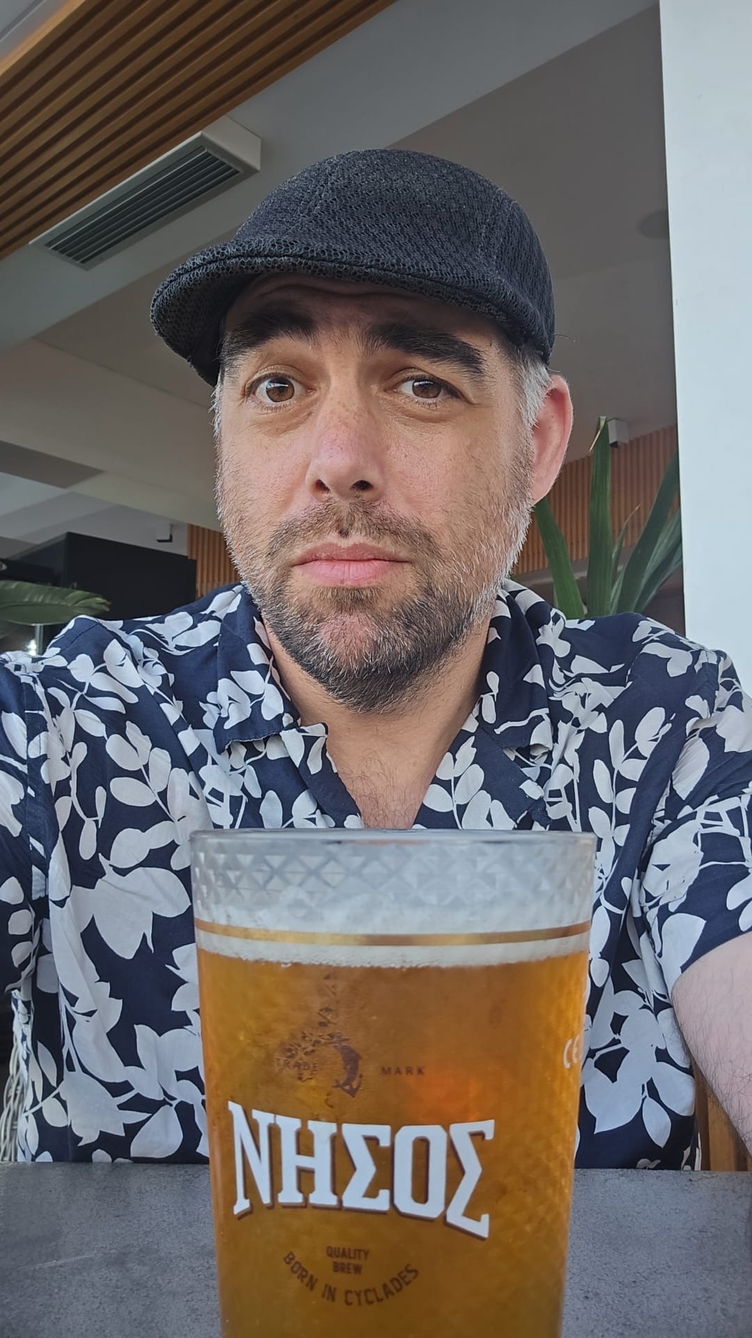 The author in a flat cap and floral shirt at a caf&eacute; in Crete, a pint of Nissos beer in the foreground reading &lsquo;Born in Cyclades.&rsquo;